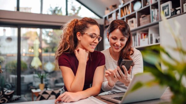 Two smiling woman looking at smart phone while sitting a desk with laptop, in modern house